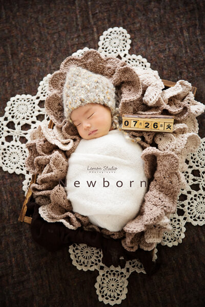 這張是一張隨手分享的很美很美的新生兒寶寶攝影相片，寶寶戴米灰色系的毛帽，包在白色的布裡，躺在大地色系及白色的花毯上，好美啊！